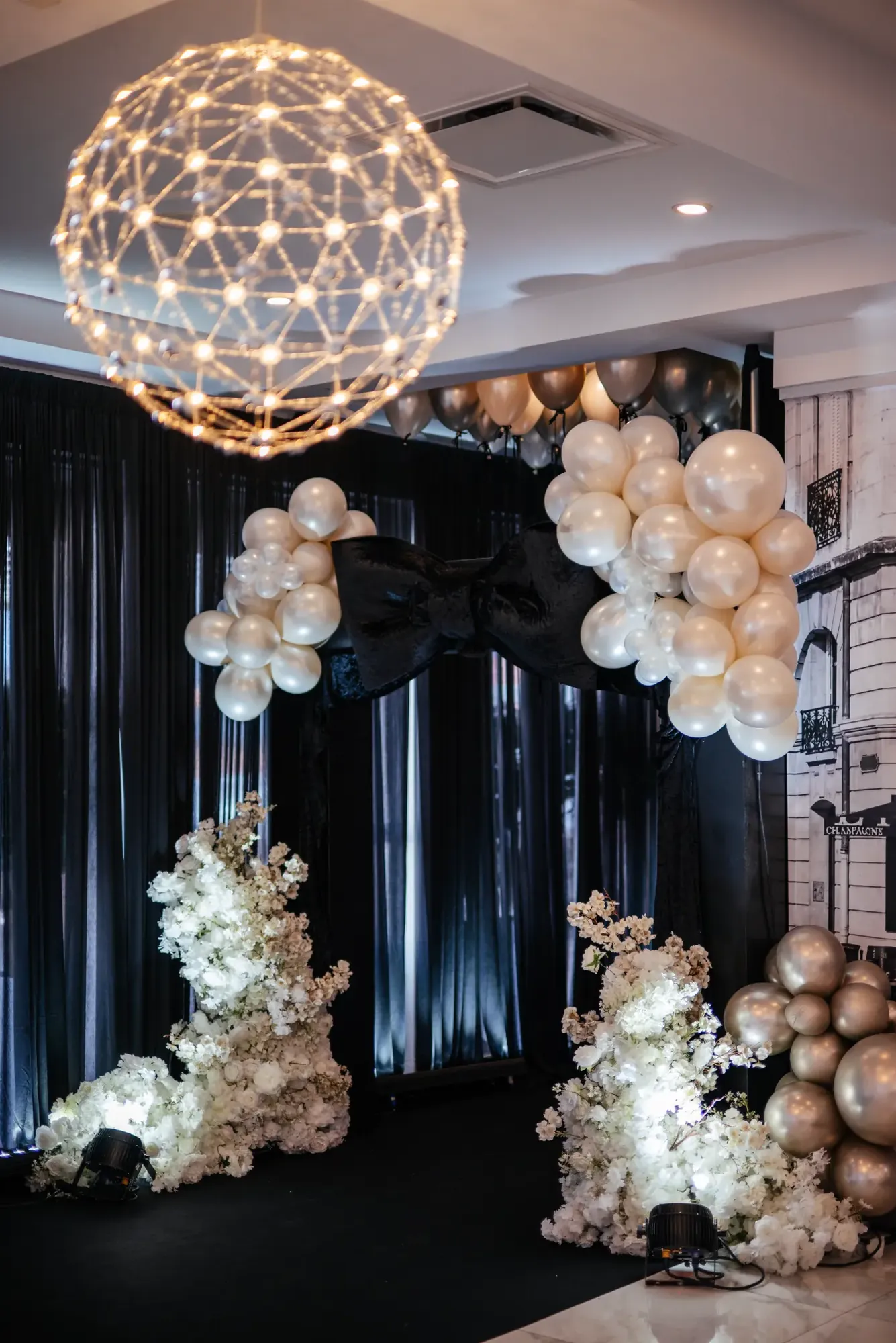 Décor de réception avec grande boucle noire, ballons blancs et arrangements floraux illuminés.